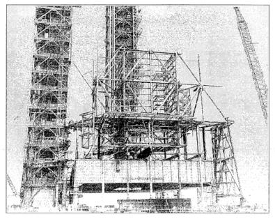 Small-format photograph: The RSS Bottom Truss Weldment, 160 feet in length, has been lifted into place at Space Shuttle Launch Complex 39-A, Kennedy Space Center, Florida, placed on Falsework which itself was place on top of an unused Apollo Program Mobile Launcher, that held it in place until the RSS became self-supporting. The Truss forms the backbone for all of the additional RSS steel which you see being erected in this photograph.